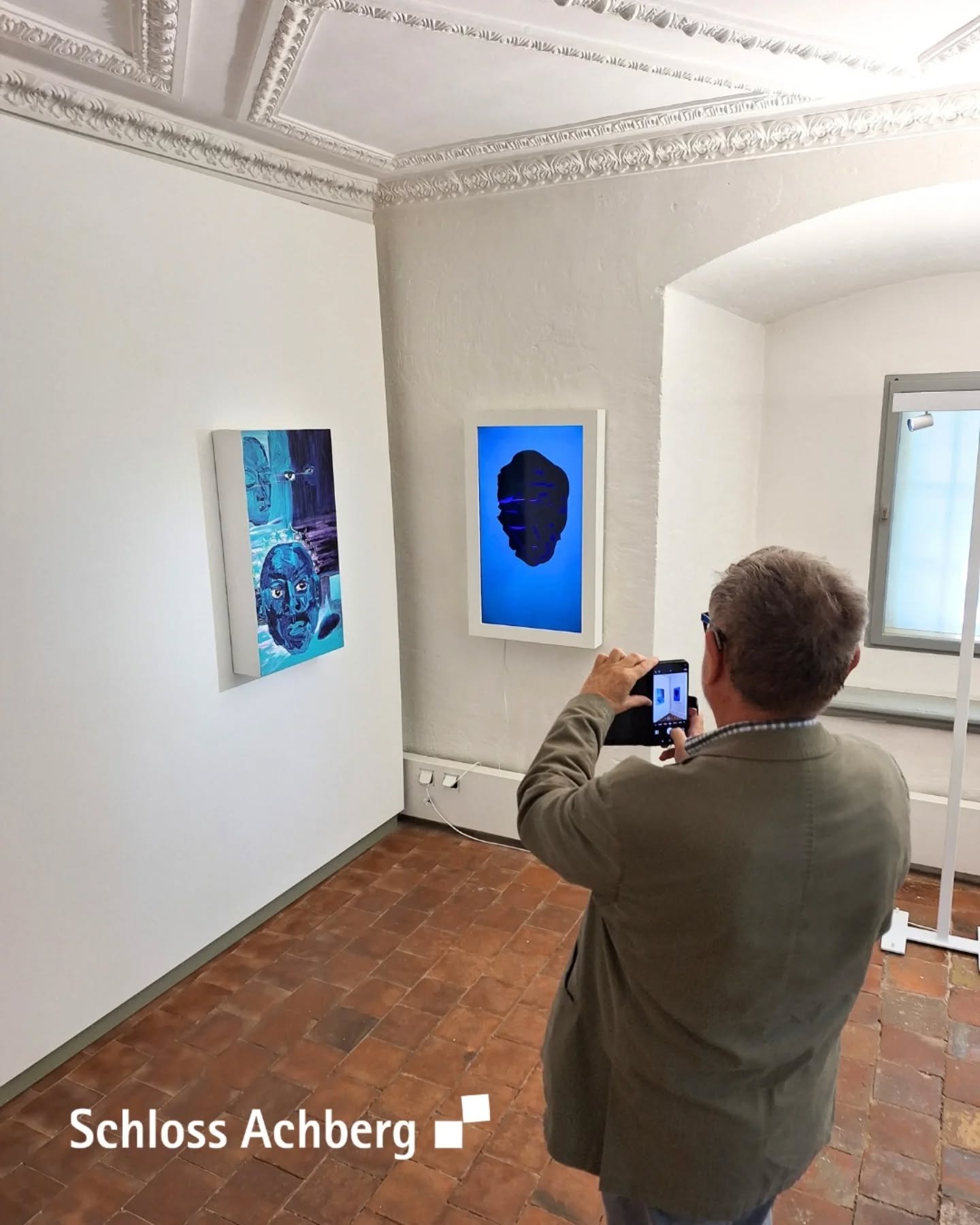 Eine Person steht in einem Raum mit Kunstwerken an den Wänden und fotografiert mit einem Mobiltelefon. Die Decke ist mit Stuck verziert.