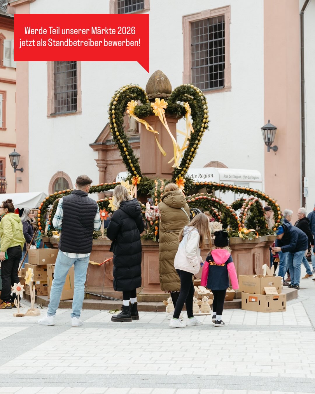 Dieser Alternativtext wurde KI-generiert: Ein Gebäude mit einer herzförmigen Dekoration aus Blumen und Früchten ist zu sehen. Mehrere Personen stehen davor und betrachten die Dekoration.
