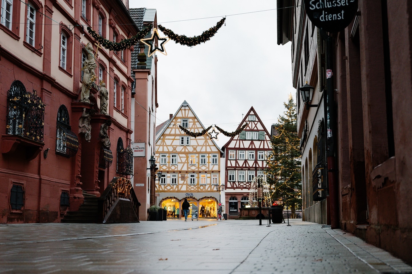 Festlich geschmückte Altstadtgasse mit Fachwerkhäusern und Weihnachtsbaum. Menschen spazieren auf dem nassen Pflaster.