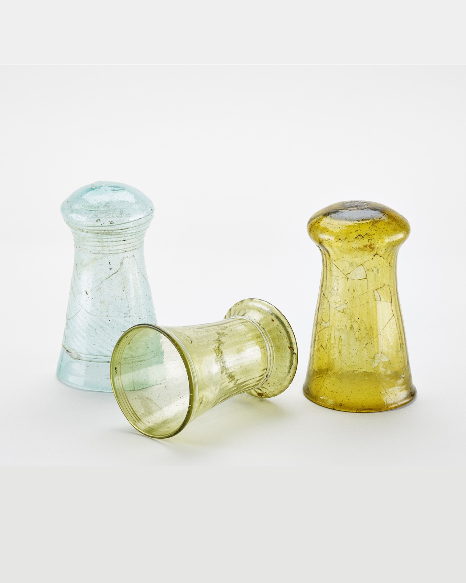 Drei kleine, alt wirkende Glasvasen oder Kerzenhalter auf weißem Hintergrund. Links steht ein hellblaues Exemplar, rechts ein gelbes, während in der Mitte ein gelbgrünes Glas auf der Seite liegt. (KI-Alternativtext)