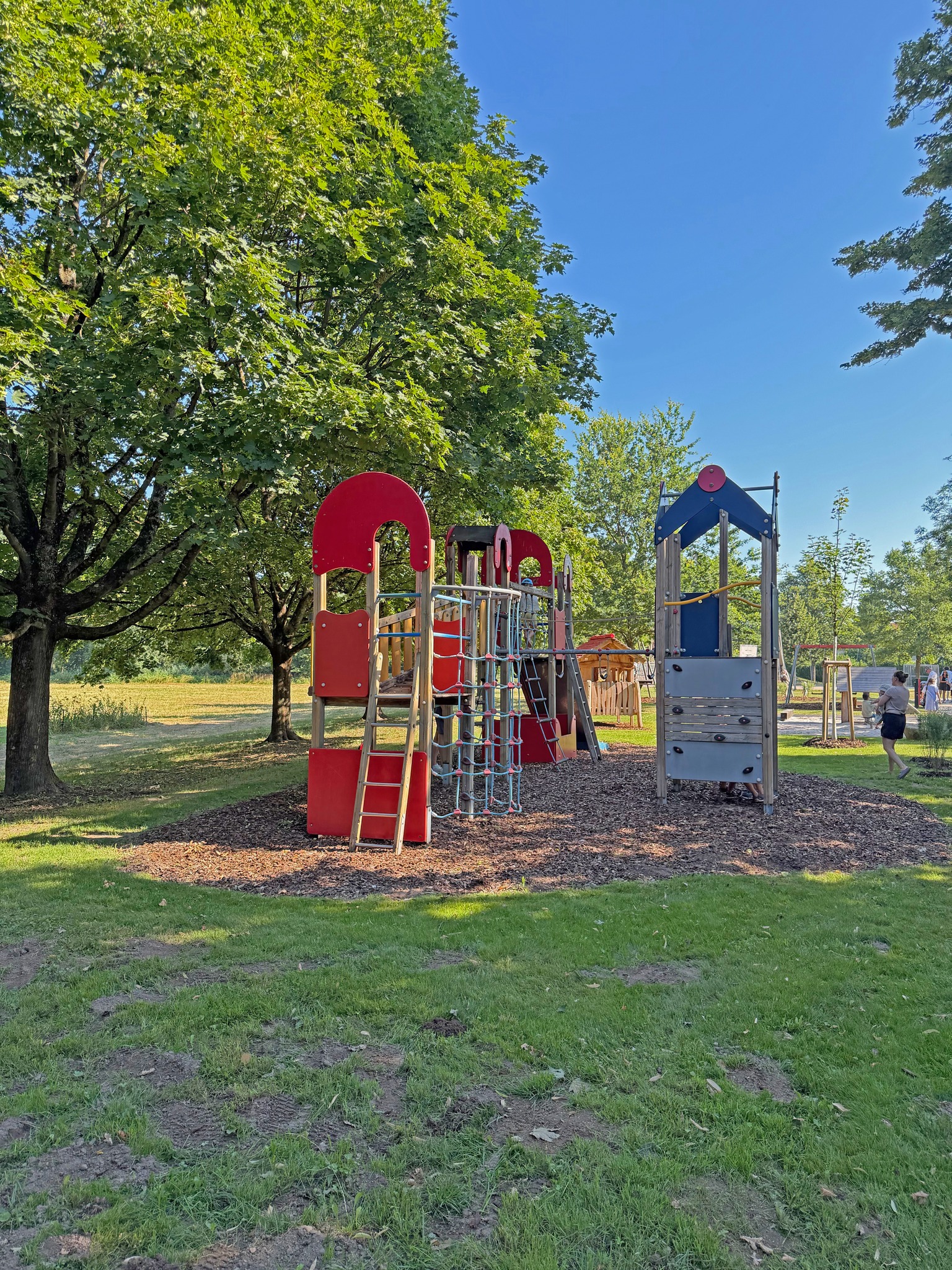 Ein Spielplatz mit hölzernen Klettergerüsten, die mit roten und blauen Paneelen, Leitern und einem Kletternetz ausgestattet sind. Die Anlage steht auf einer Fläche mit Holzspänen, umgeben von Gras und großen Bäumen unter blauem Himmel. Rechts im Hintergrund läuft eine Person vorbei. (KI-Alternativtext)