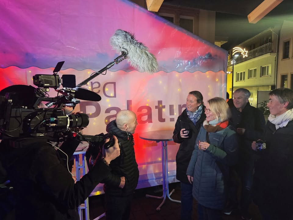 Dieser Alternativtext wurde KI-generiert: Eine Filmkamera mit Mikrofon ist vor einer beleuchteten Bühnenkulisse aufgebaut. Mehrere Personen stehen im Hintergrund und beobachten die Aufnahme.