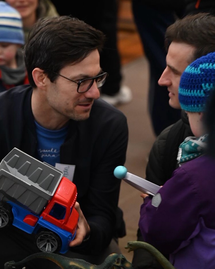 Dieser Alternativtext wurde KI-generiert: Zwei Personen unterhalten sich, während ein Kind mit Spielzeugautos spielt. Das Bild ist farbig und zeigt eine entspannte Interaktion.