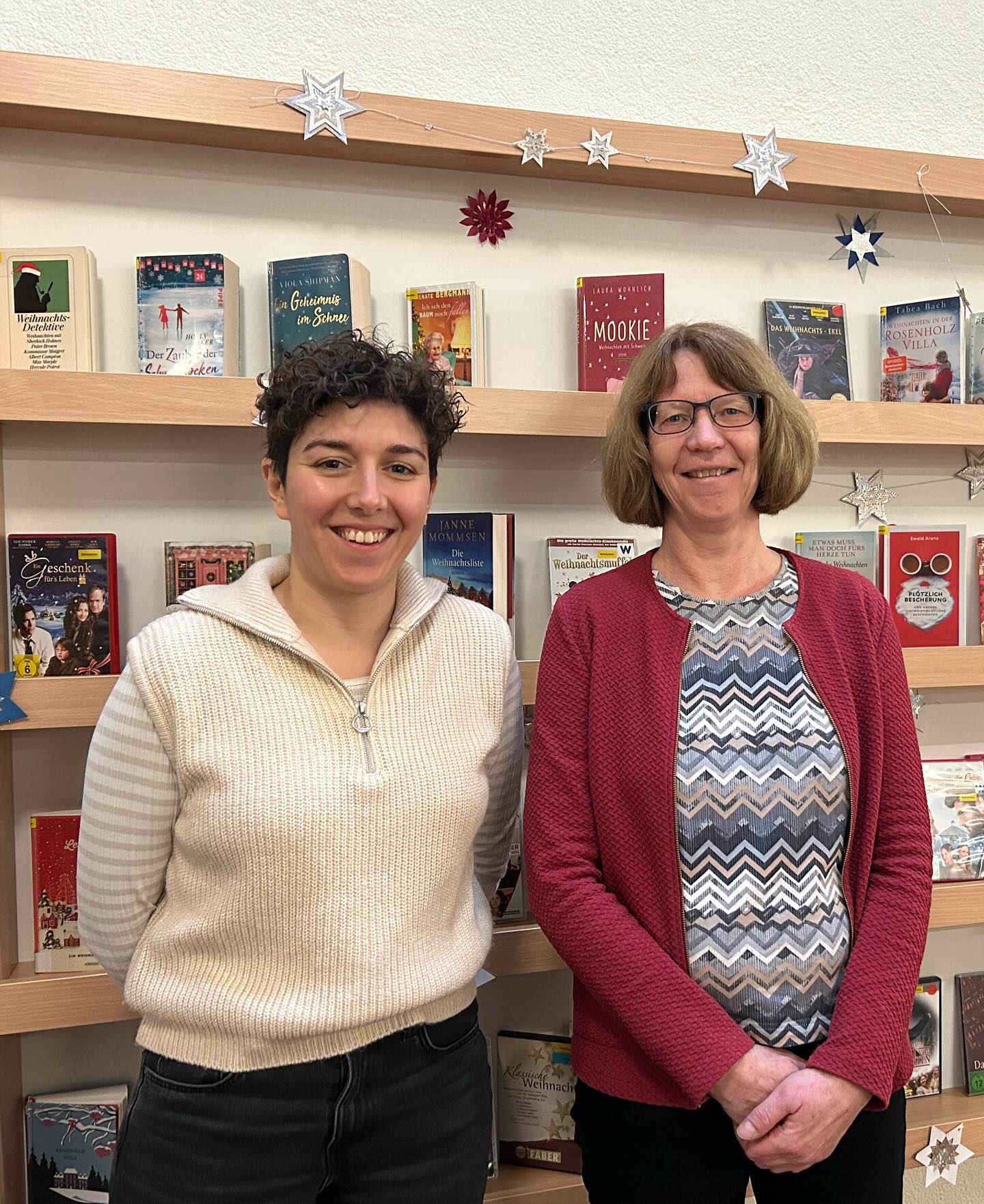 Dieser Alternativtext wurde KI-generiert: Zwei Personen stehen vor einem Bücherregal mit weihnachtlichen Büchern. Das Bild ist farbig und zeigt eine gemütliche Atmosphäre.