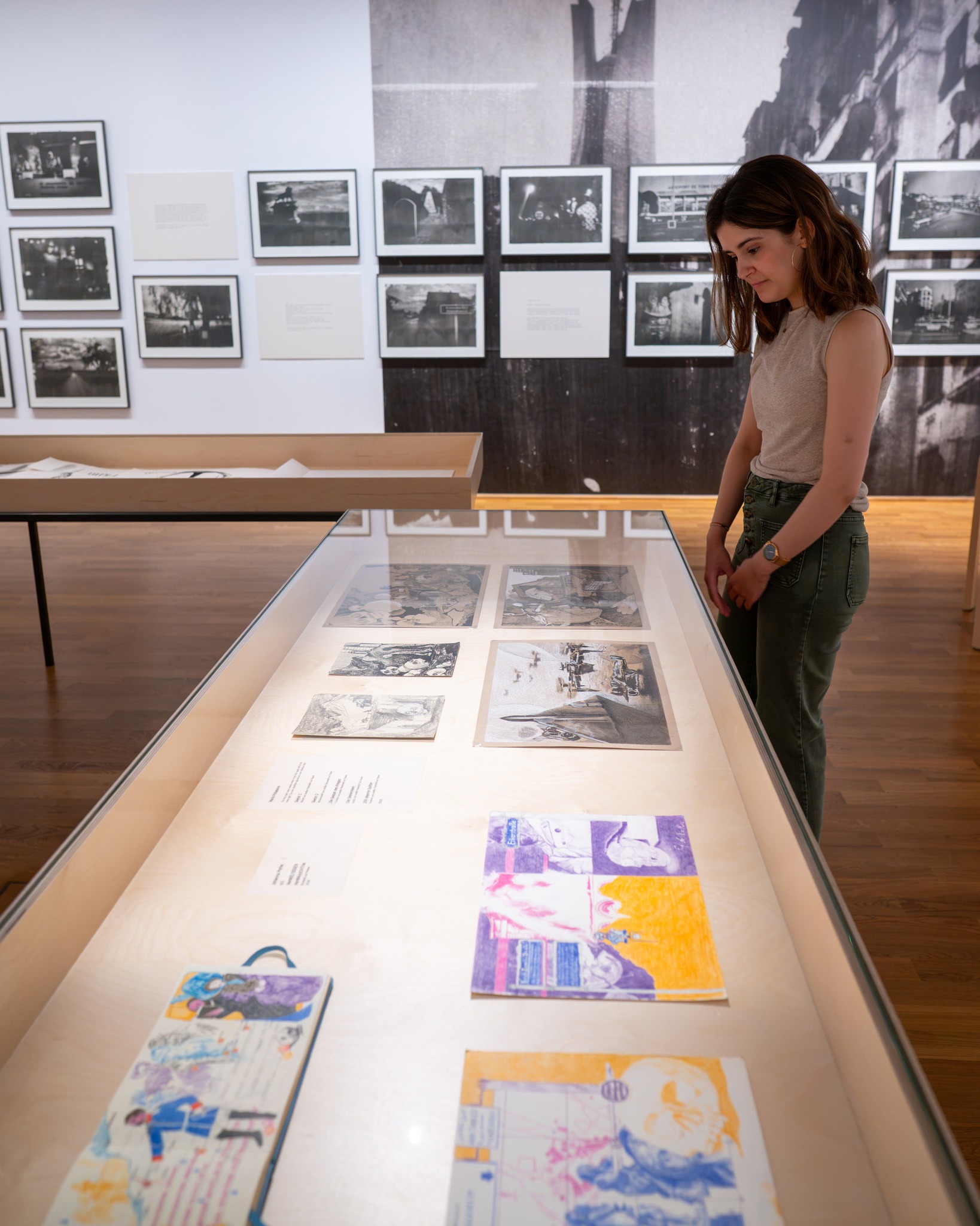 Eine Person in einem ärmellosen Oberteil und grüner Hose betrachtet in einer Ausstellung verschiedene Dokumente und Zeichnungen in einer langen Glasvitrine. An der Wand dahinter sind gerahmte Schwarz-Weiß-Fotografien und Texttafeln zu sehen. (KI-Alternativtext)