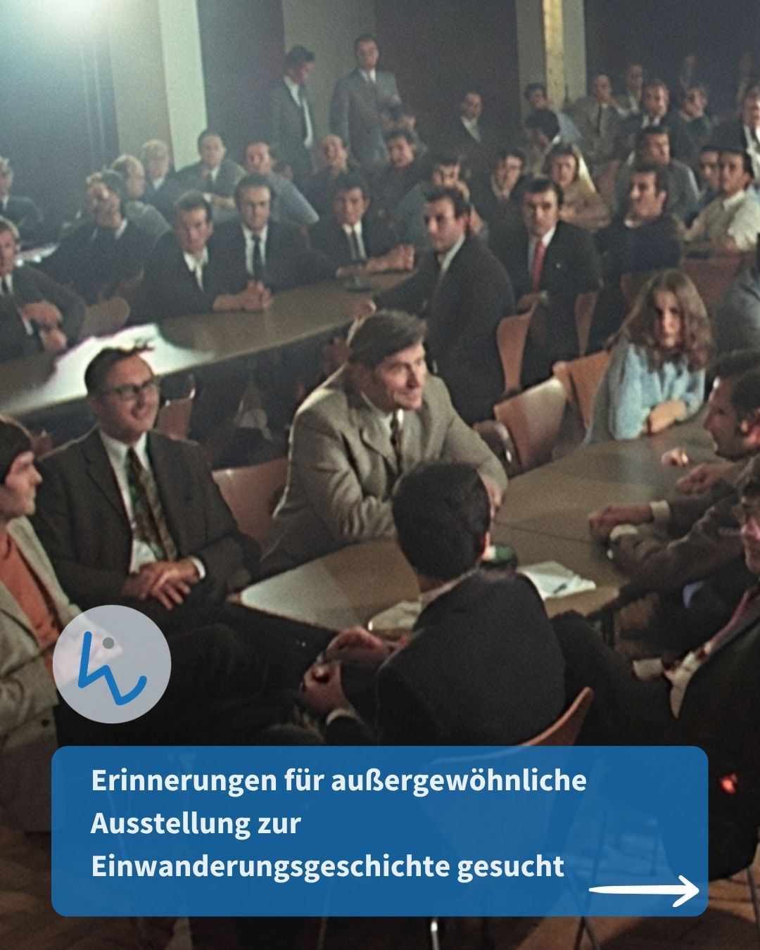Eine große Gruppe von Personen in formeller Kleidung sitzt an langen Tischen in einem Saal und nimmt an einer Versammlung teil. Ein blauer Balken unten enthält den Text: "Erinnerungen für außergewöhnliche Ausstellung zur Einwanderungsgeschichte gesucht" sowie einen Pfeil nach rechts. (KI-Alternativtext)