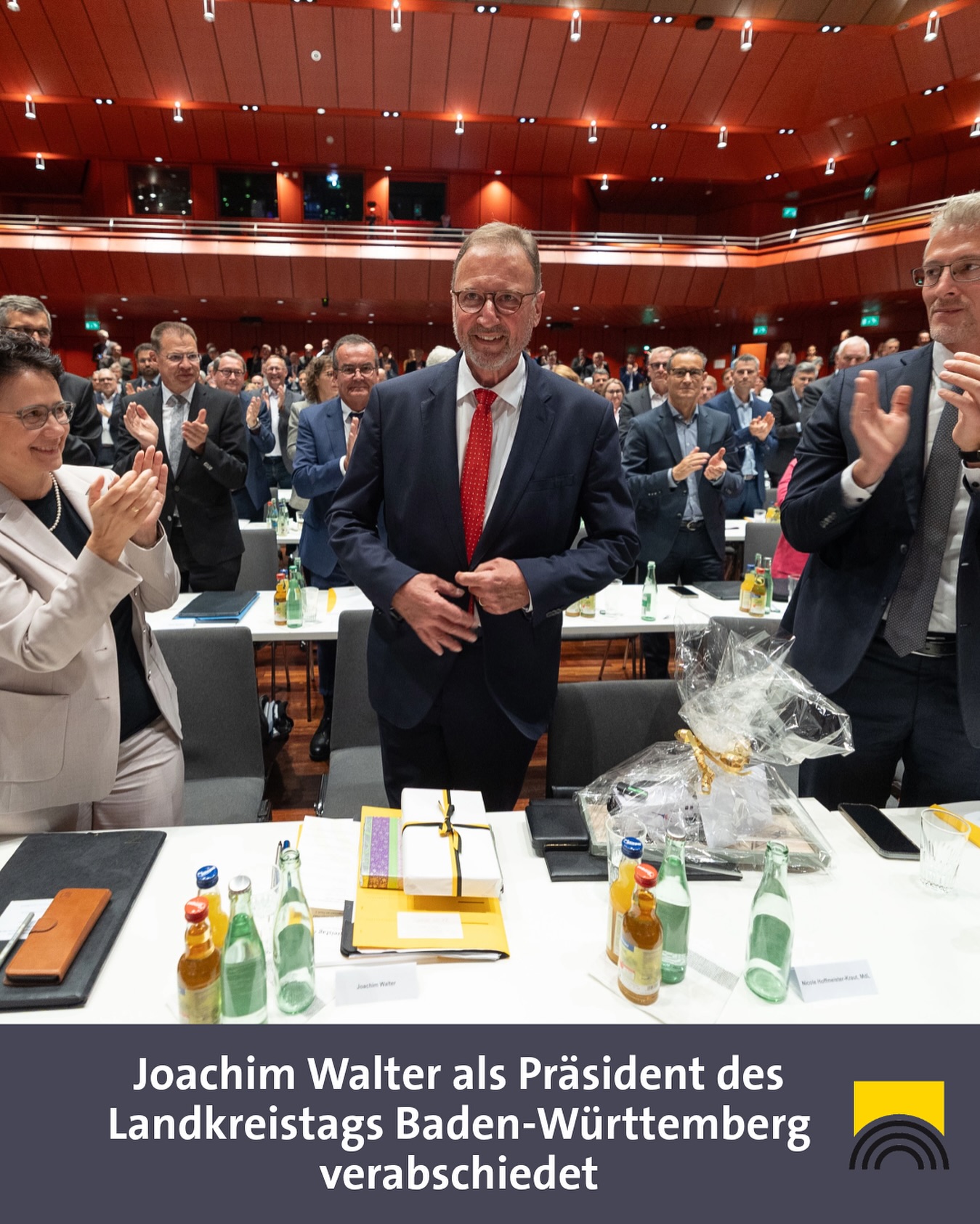 Dieser Alternativtext wurde KI-generiert: Eine Innenaufnahme eines großen Konferenzraums mit vielen Sitzenden. Im Vordergrund steht eine Person, die freundlich in die Kamera lächelt und von anderen umgeben ist.