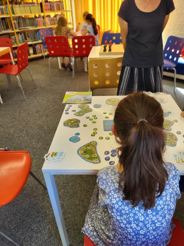 Eine Person mit langem Haar sitzt an einem weißen Tisch und spielt ein Brettspiel mit grünen und blauen Landschaftssteinen sowie kleinen Figuren. Eine weitere Person steht hinter dem Tisch. Im Hintergrund sind Bücherregale und weitere Personen an Tischen zu sehen. (KI-Alternativtext)