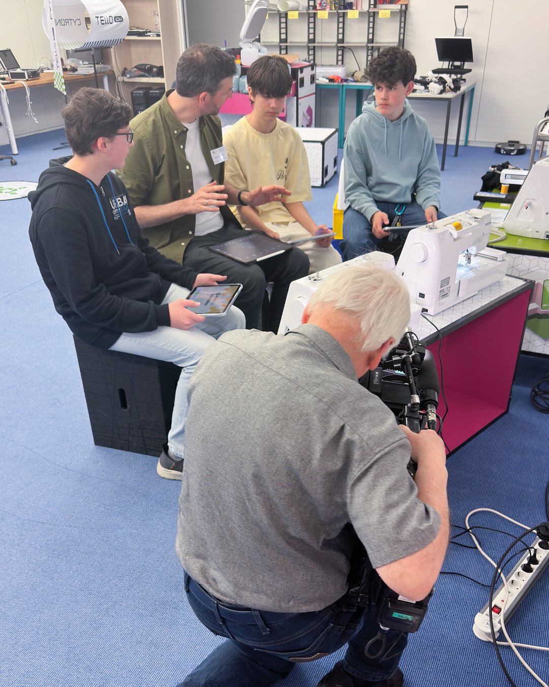 Dieser Alternativtext wurde KI-generiert: Eine Gruppe von Personen ist in einem Raum mit technischer Ausrüstung zu sehen. Eine Person kniet und filmt die anderen mit einer Kamera.