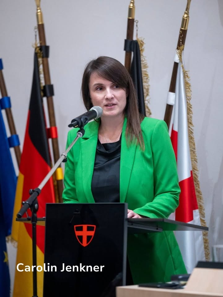 Dieser Alternativtext wurde KI-generiert: Eine Person steht an einem Rednerpult und spricht in ein Mikrofon. Im Hintergrund sind mehrere Flaggen zu sehen.