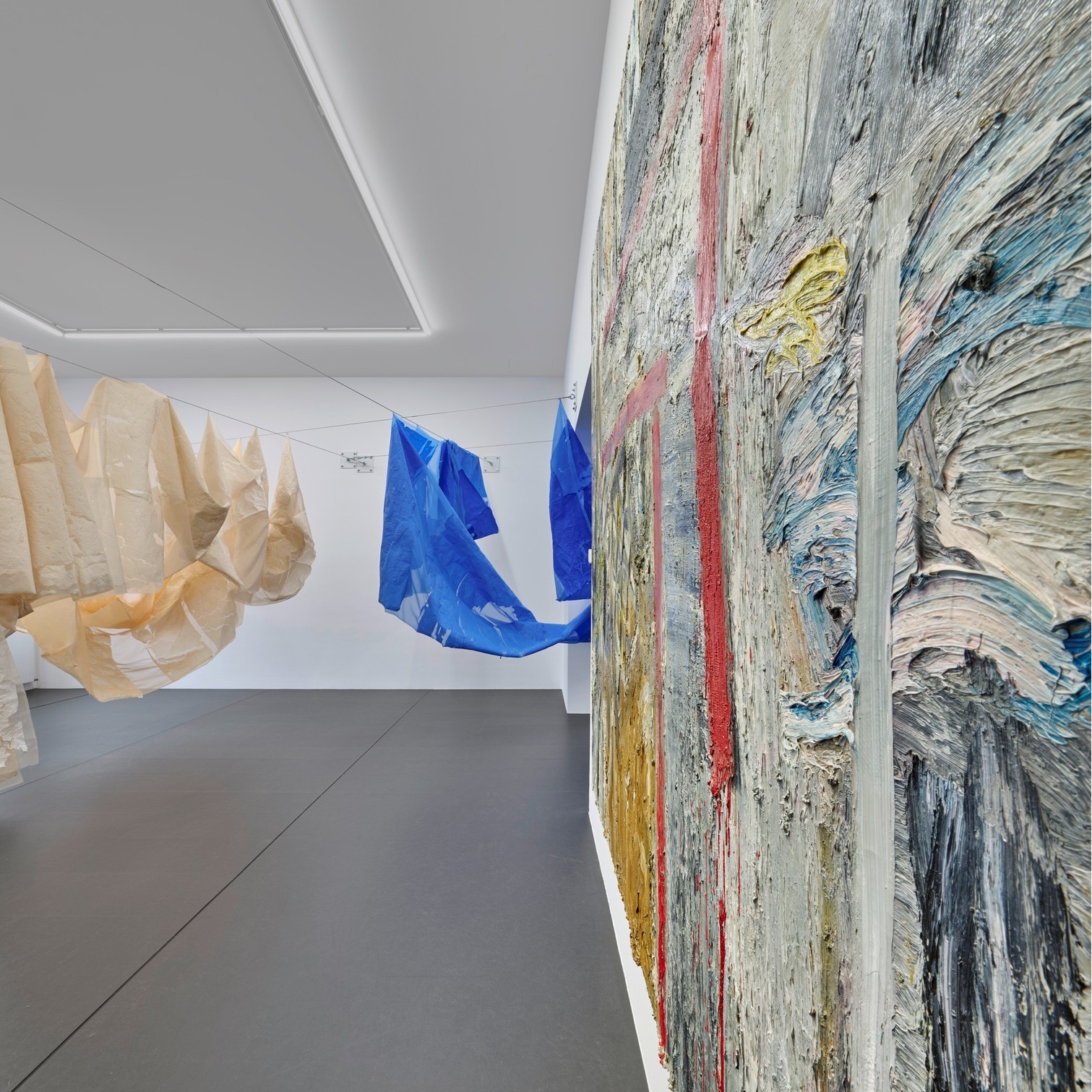 Dieser Alternativtext wurde KI-generiert: Eine Galerieansicht mit einem farbenfrohen, strukturierten Gemälde an der Wand und hängenden Stoffstrukturen im Raum. Der Raum hat einen hellen Boden und eine helle Decke mit eingebauter Beleuchtung.