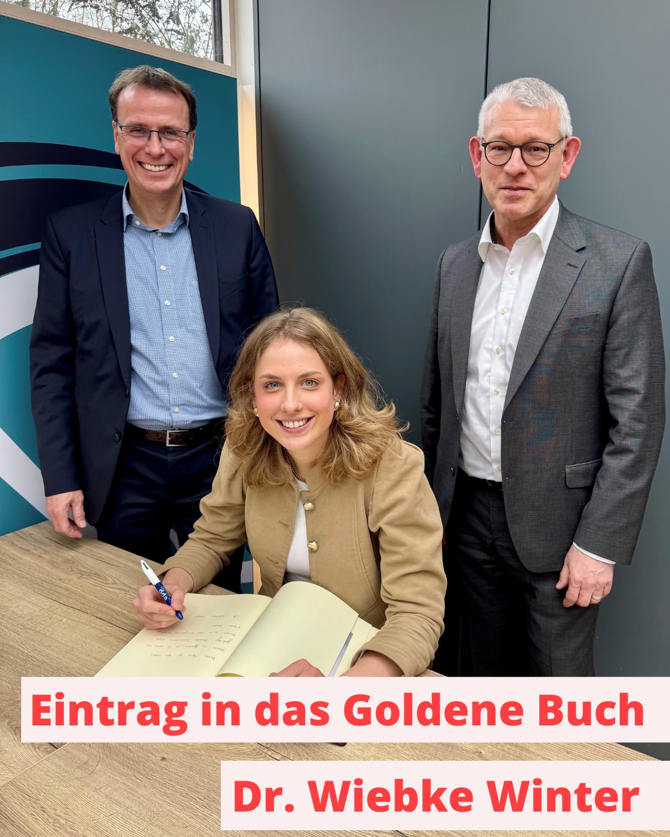 Dieser Alternativtext wurde KI-generiert: Drei Personen stehen vor einem Fenster und blicken auf ein offenes Buch. Es handelt sich um eine Farbfotografie mit roter Beschriftung im unteren Bildbereich.