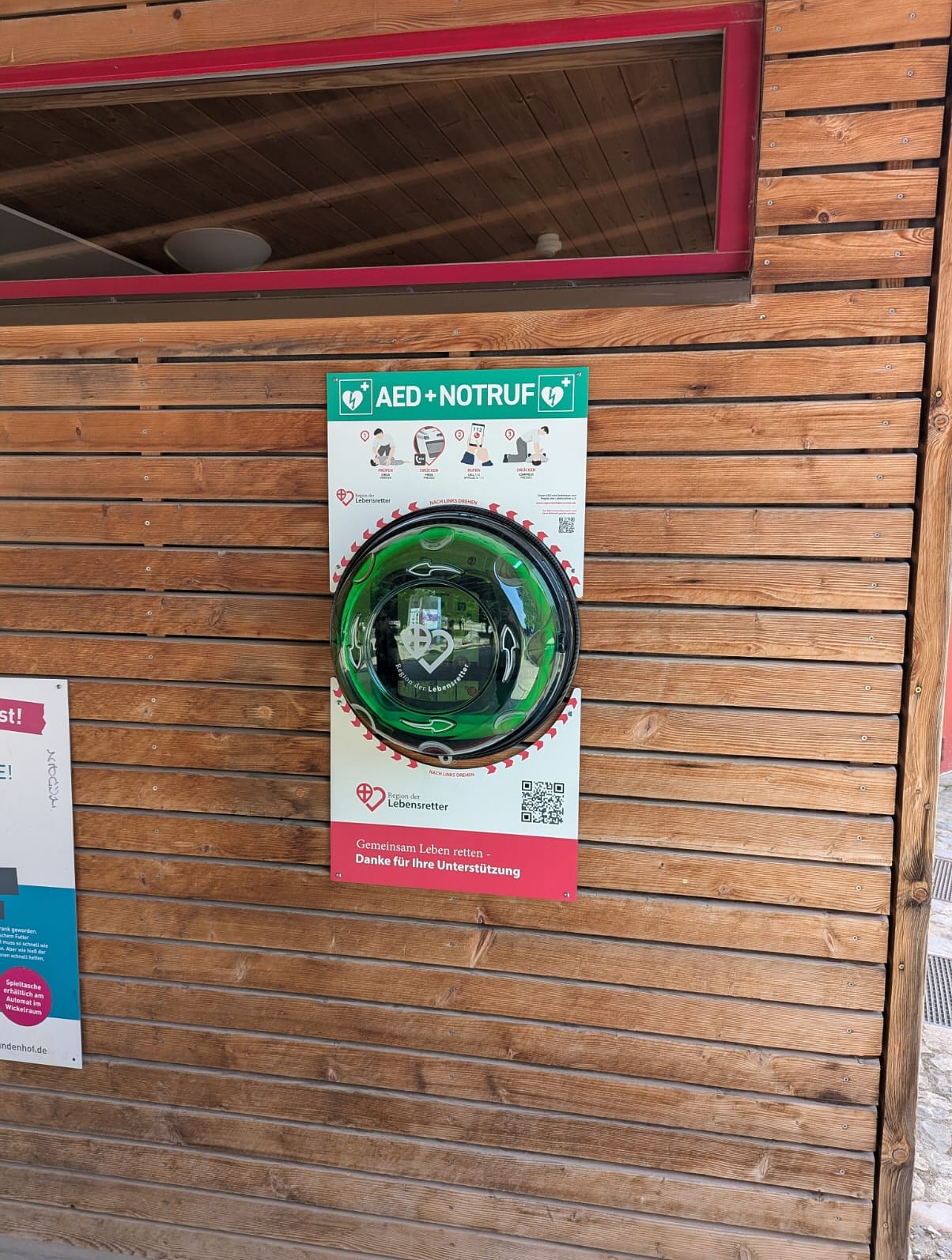 An einer Holzwand ist ein AED-Defibrillator mit zugehöriger Anleitung befestigt. Das Gerät befindet sich in einem runden Behälter.