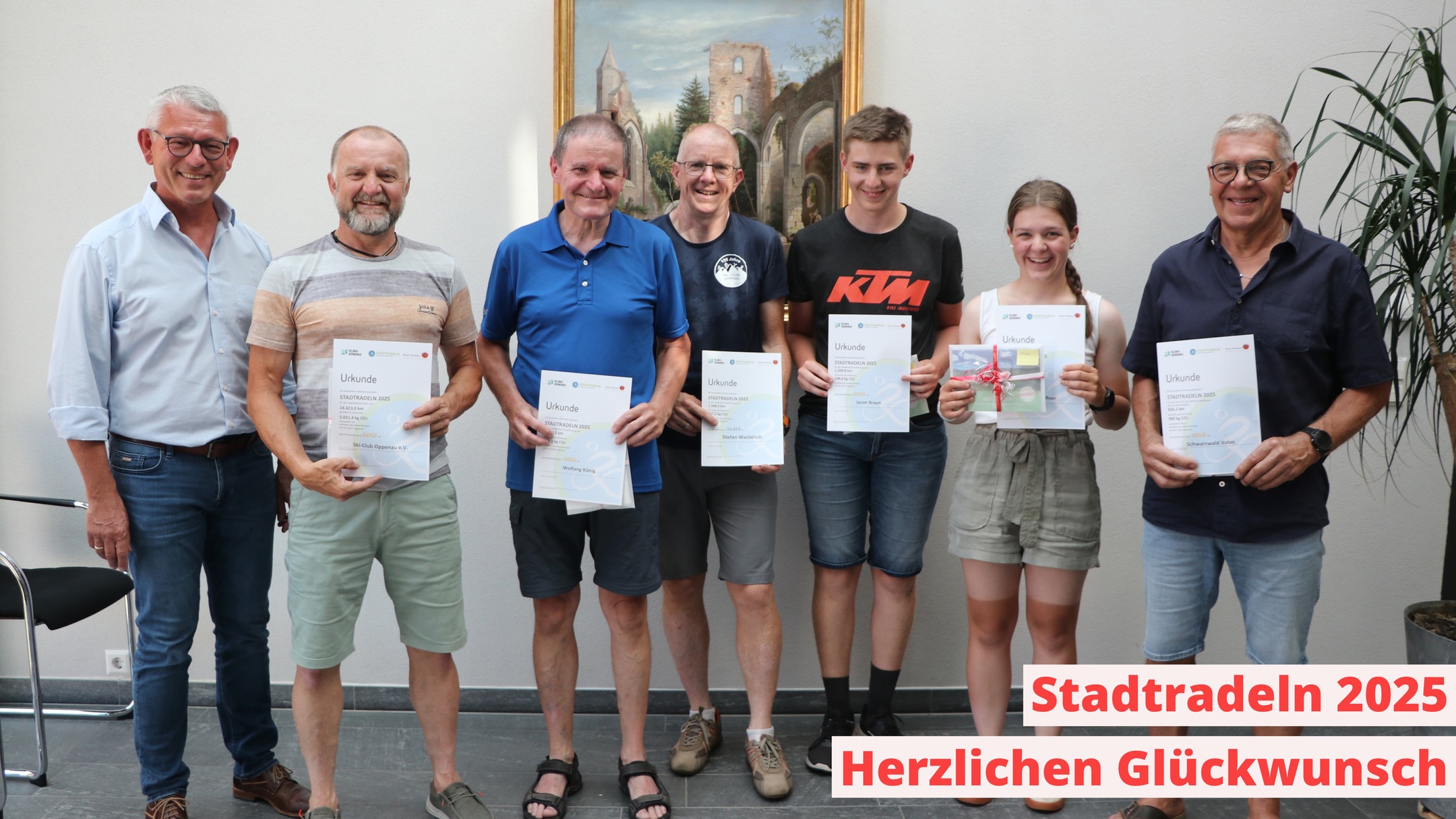 Eine Gruppe hält Urkunden. Im Hintergrund hängt ein gerahmtes Gemälde und eine Zimmerpflanze steht in der Ecke.