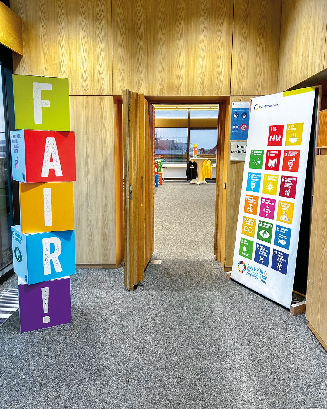 Dieser Alternativtext wurde KI-generiert: Ein Innenraum zeigt eine Türöffnung mit Blick auf einen weiteren Raum. Vor der Tür steht ein farbenfroher Turm aus Buchstabenwürfeln und ein Poster mit Symbolen.