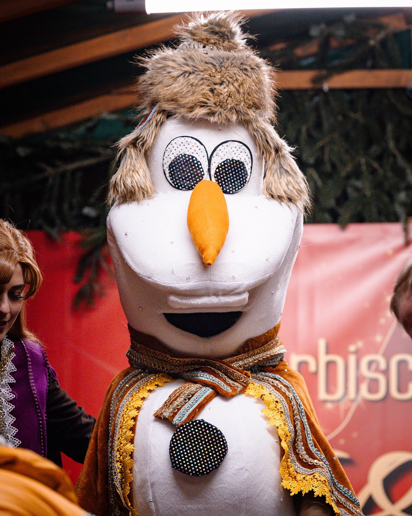 Ein niedliches Schneemannkostüm mit einem freundlichen Gesicht und einem braunen Pelzbesatz. Im Hintergrund sind weitere Personen zu sehen.