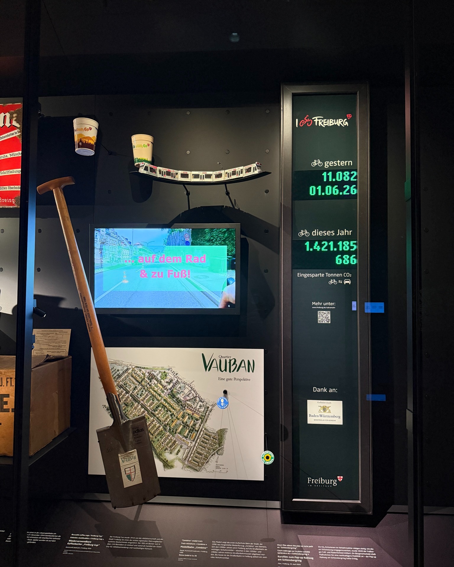 Ausstellungsvitrine mit Spaten, Modellbahn, Bildschirm mit Text „… auf dem Rad & zu Fuß!“, Stadtplan des Quartiers Vauban und digitaler Anzeige für Fahrradkilometer und eingesparte CO2-Tonnen in Freiburg. (KI-Alternativtext)