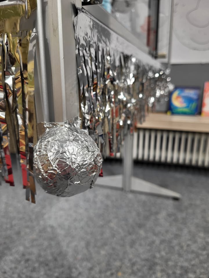 Dieser Alternativtext wurde KI-generiert: Eine Nahaufnahme zeigt glänzende, metallisch wirkende Dekorationen, die an einem Rahmen hängen. Im Hintergrund ist ein Raum mit Möbeln und Gegenständen zu erkennen.