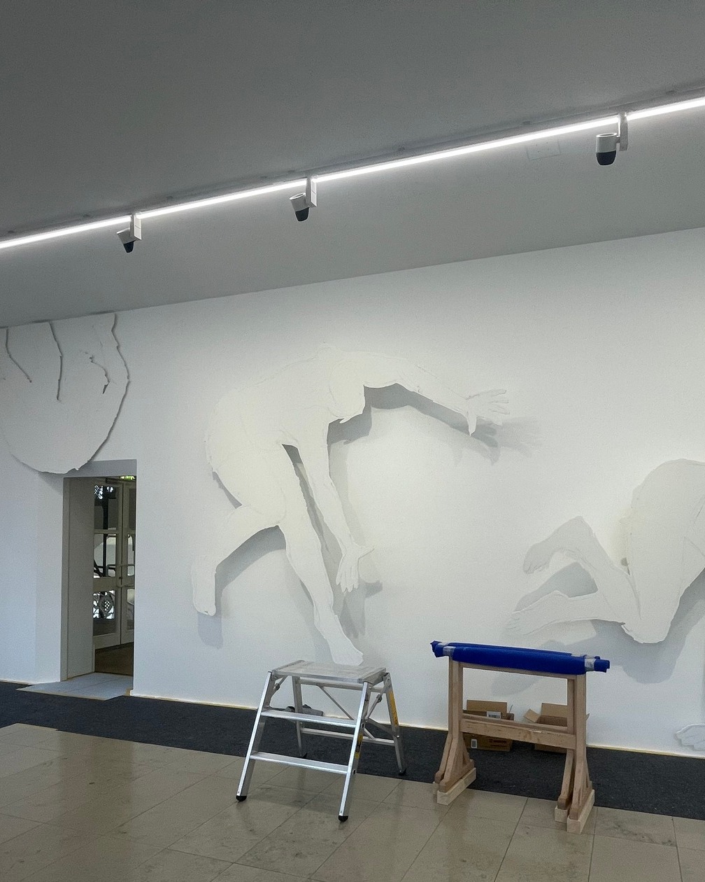 Dieser Alternativtext wurde KI-generiert: Ein Innenraum mit einer hellen Wand und einem Relief von Tierfiguren. Vor der Wand stehen eine Leiter und eine Rolle mit Material.