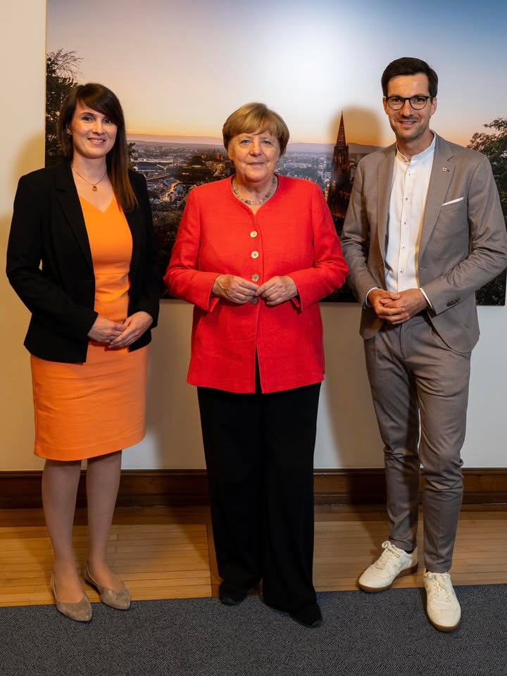 Drei Personen stehen nebeneinander vor einer Wand mit Stadtbild im Hintergrund. Die mittlere Person trägt eine rote Jacke, die linke ein orangefarbenes Kleid mit schwarzer Jacke, die rechte einen grauen Anzug mit Brille. Alle blicken in die Kamera. (KI-Alternativtext)