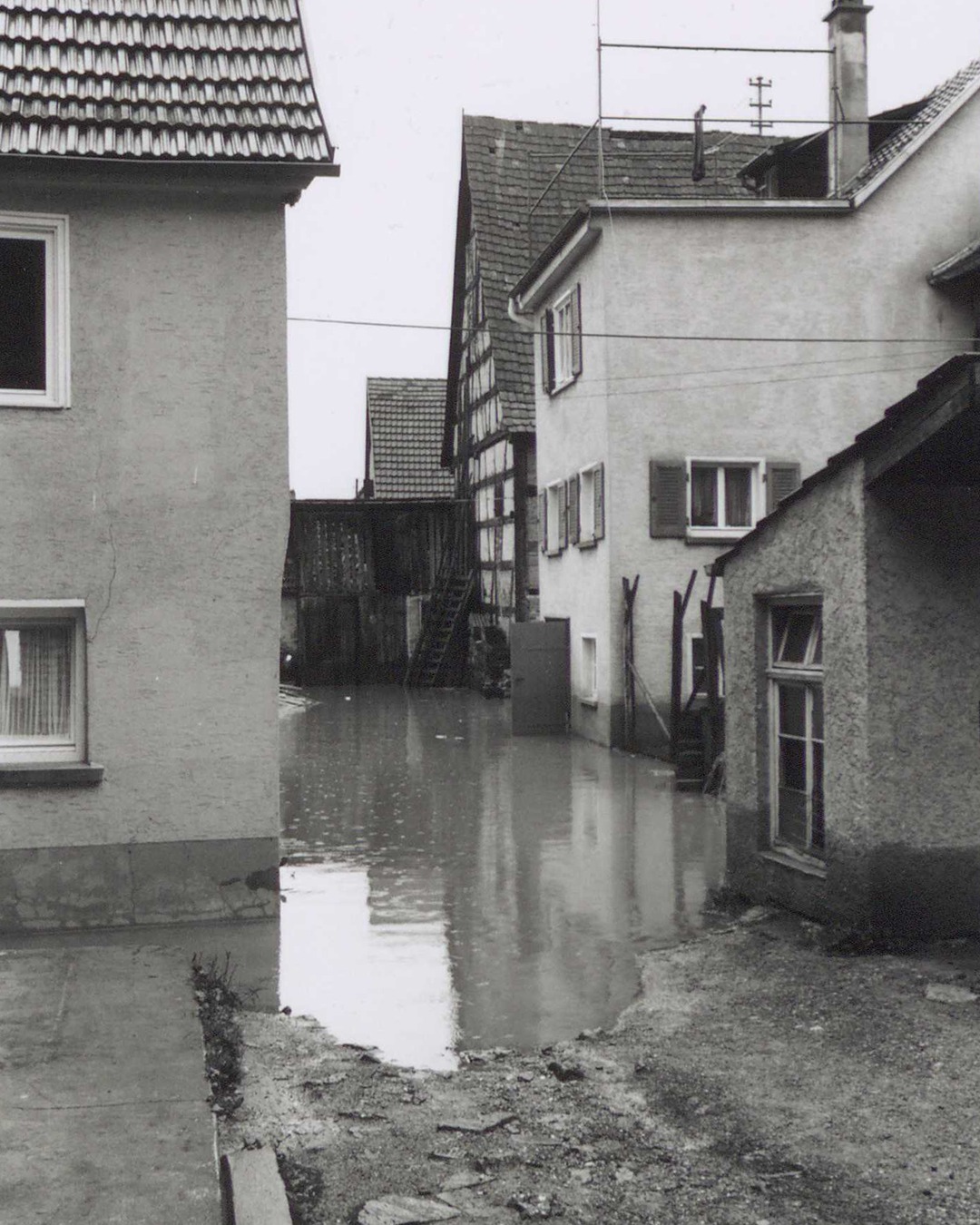Schwarz-Weiß-Foto einer überfluteten Straße zwischen mehreren Häusern unterschiedlicher Architektur mit Ziegeldächern und Fachwerk. Das Wasser bedeckt den gesamten Boden bis zu den Fundamenten der Gebäude und reflektiert deren Fassaden im Hintergrund. (KI-Alternativtext)