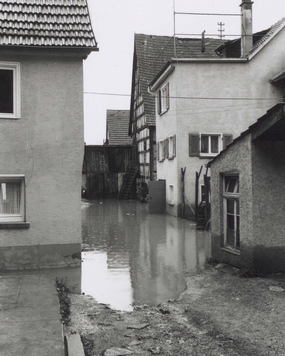 Schwarz-Weiß-Foto einer überfluteten Straße zwischen mehreren Häusern unterschiedlicher Architektur mit Ziegeldächern und Fachwerk. Das Wasser bedeckt den gesamten Boden bis zu den Fundamenten der Gebäude und reflektiert deren Fassaden im Hintergrund. (KI-Alternativtext)