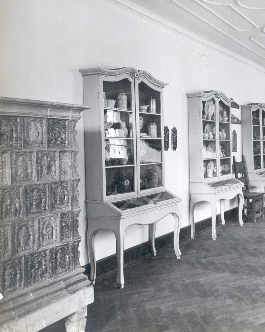 Dieser Alternativtext wurde KI-generiert: Schwarz-weißes Bild eines Innenraums mit mehreren alten Vitrinenschränken an einer Wand. Ein Teil eines steinernen Kamins ist ebenfalls zu sehen.