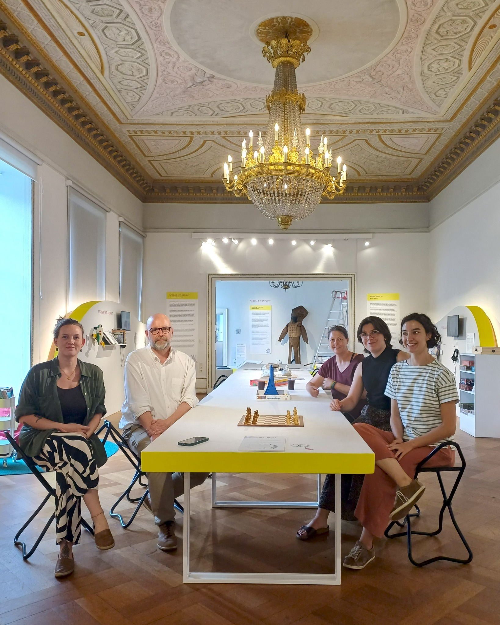 Fünf Personen sitzen um einen langen weißen Tisch mit gelbem Rand in einem Ausstellungsraum mit verziertem Deckengewölbe und großer Kronleuchter. Auf dem Tisch befinden sich ein Schachbrett und weitere Spielobjekte. An den Wänden sind Infotafeln angebracht, darunter eine mit der Aufschrift „SPIELEN MIT ABSICHT“. (KI-Alternativtext)