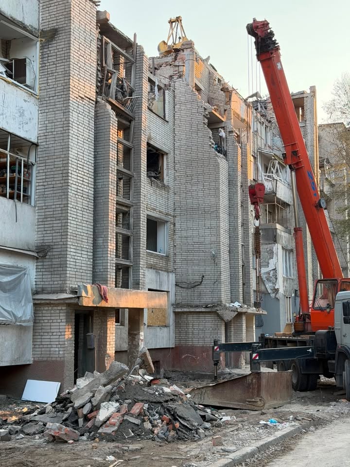 Mehrstöckiges Backsteingebäude mit stark beschädigter Fassade und fehlenden Fenstern im Abrisszustand. Rechts positioniert sich ein großer orangefarbener Kran, dessen Arm in Richtung des Gebäudes zeigt. Im Vordergrund liegt ein großer Haufen aus Ziegel- und Betontrümmern. (KI-Alternativtext)