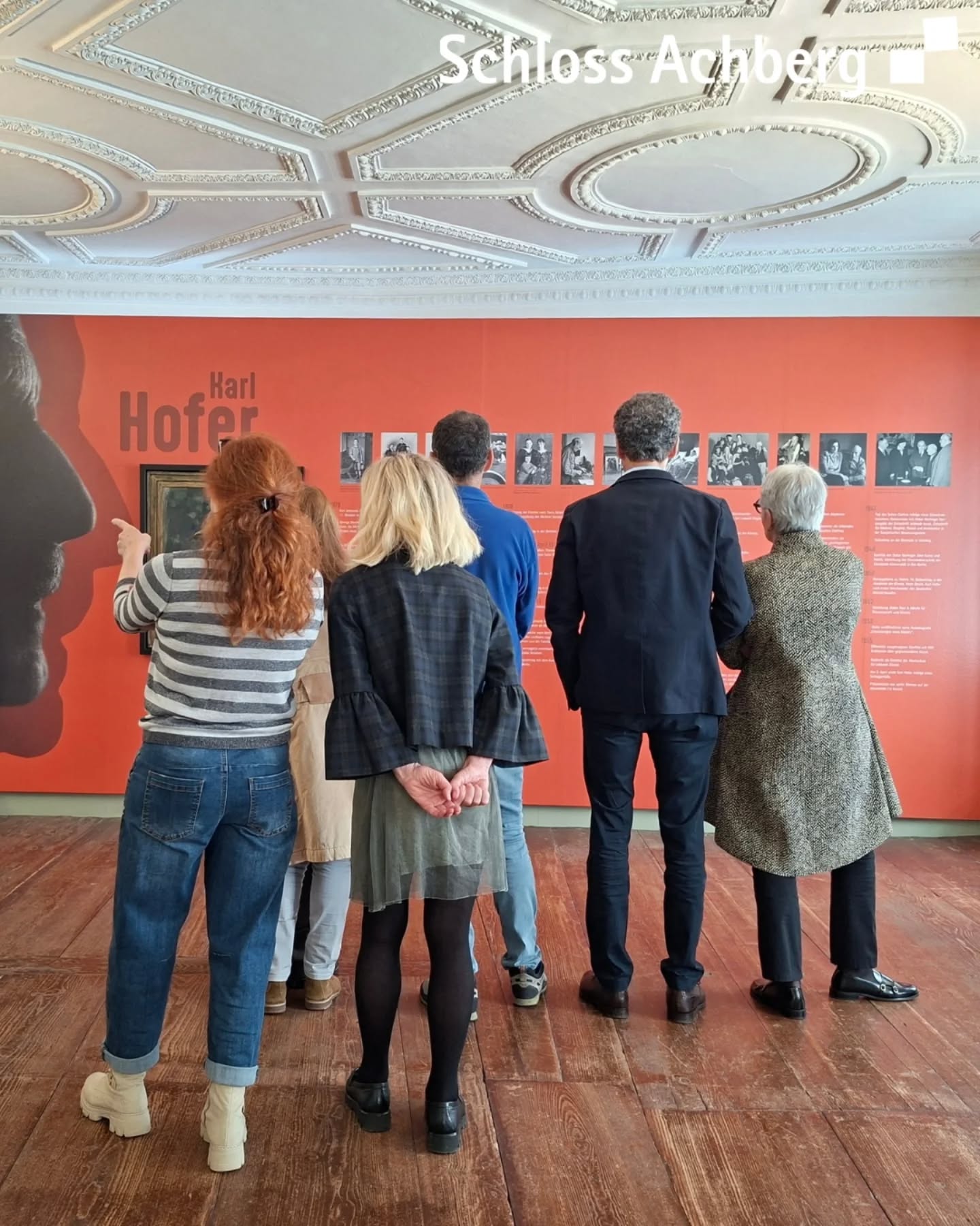 Dieser Alternativtext wurde KI-generiert: Eine Gruppe von Menschen betrachtet in einem Raum eine Ausstellung mit Fotografien an einer Wand. Der Raum hat eine stuckverzierte Decke und einen Holzboden.