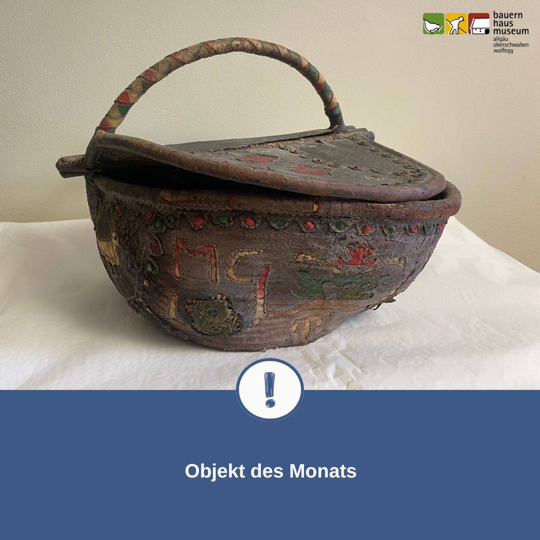 Alter, verzierter Korb mit Deckel und Henkel, bemalt mit Buchstaben und Mustern, präsentiert als „Objekt des Monats“ vom Bauernhausmuseum. (KI-Alternativtext)