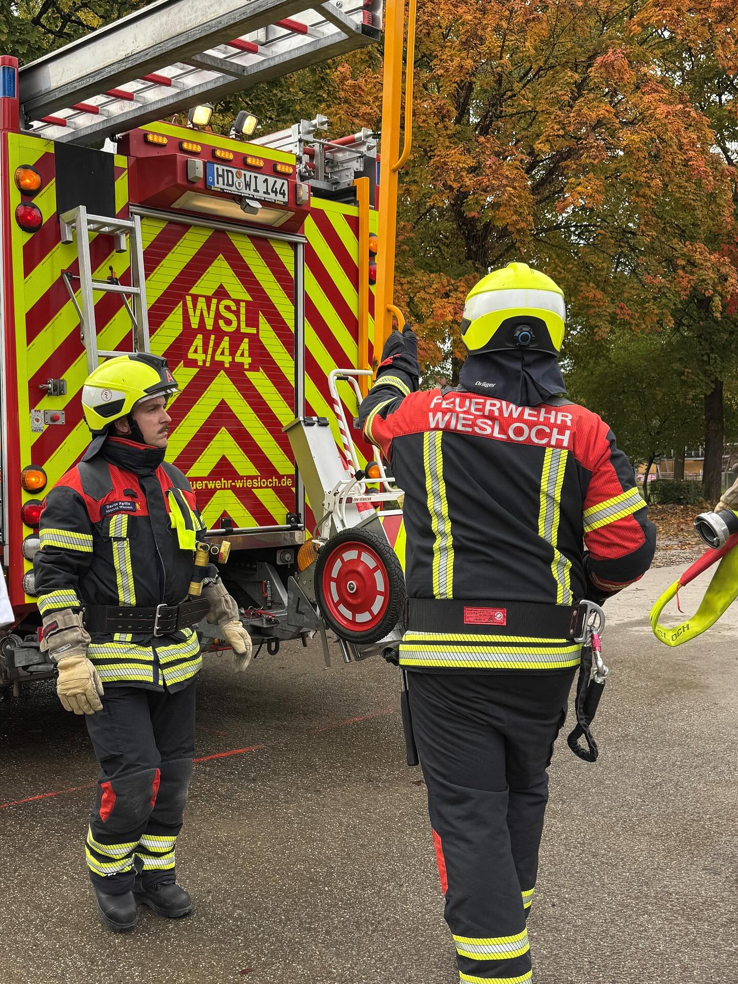 Dieser Alternativtext wurde KI-generiert: Ein rotes Fahrzeug mit gelben Streifen steht im Hintergrund. Personen in schwarzer und roter Einsatzkleidung stehen davor, eine Person blickt in die Kamera.