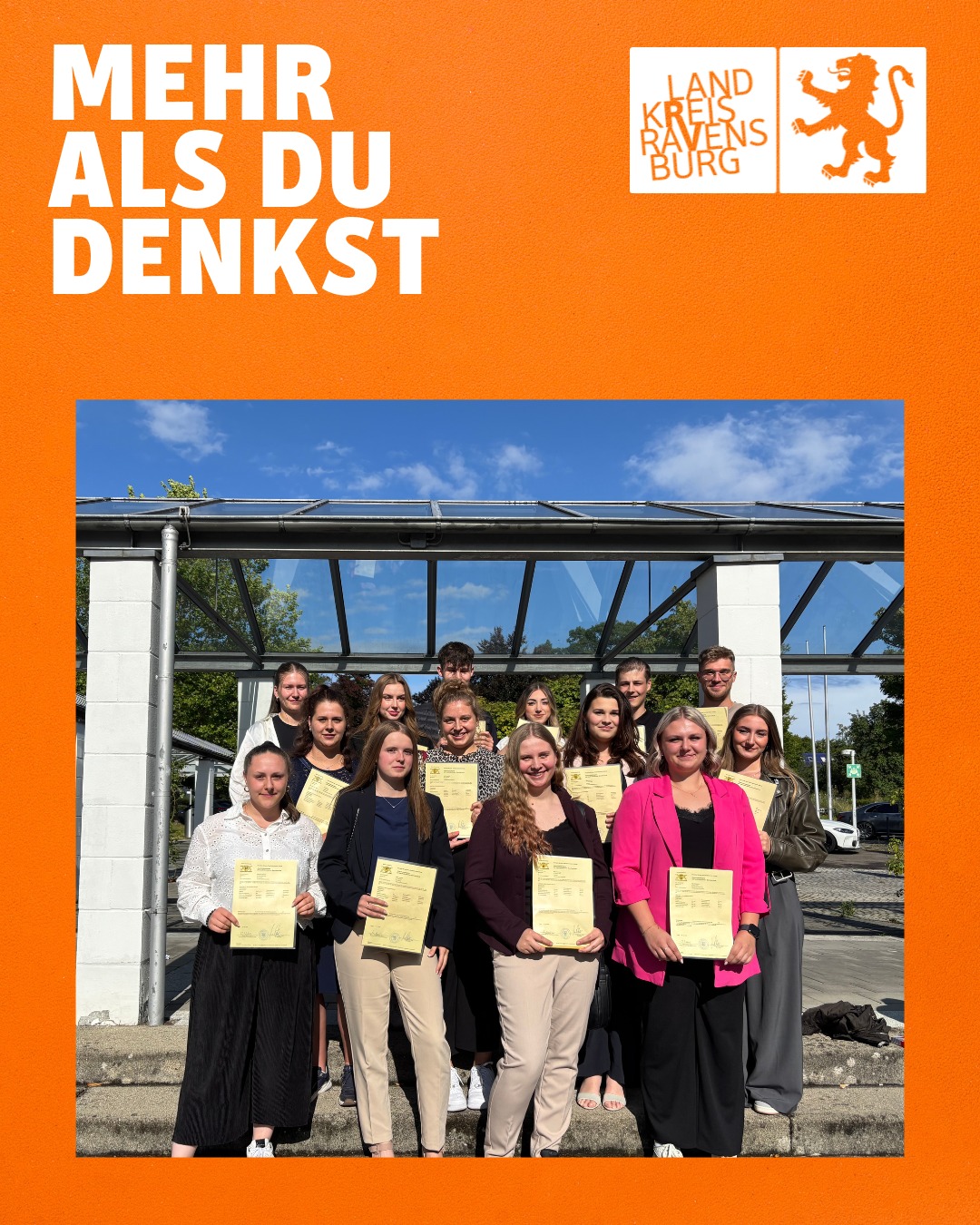 Eine Gruppe von Personen steht vor einem Gebäude und hält Urkunden. Das Bild hat einen orangenen Rahmen mit Text und einem Logo.
