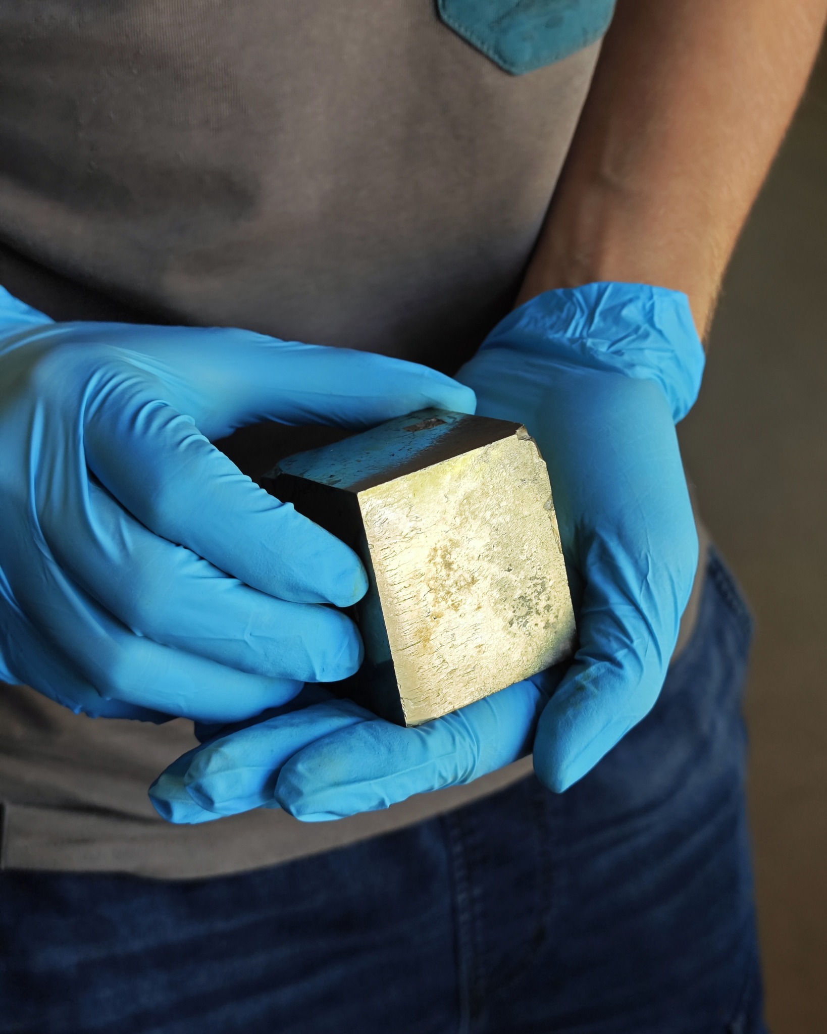 Zwei Hände in blauen Einweghandschuhen halten einen rechteckigen Goldbarren. Die Person trägt ein graues T-Shirt und blaue Jeans. (KI-Alternativtext)