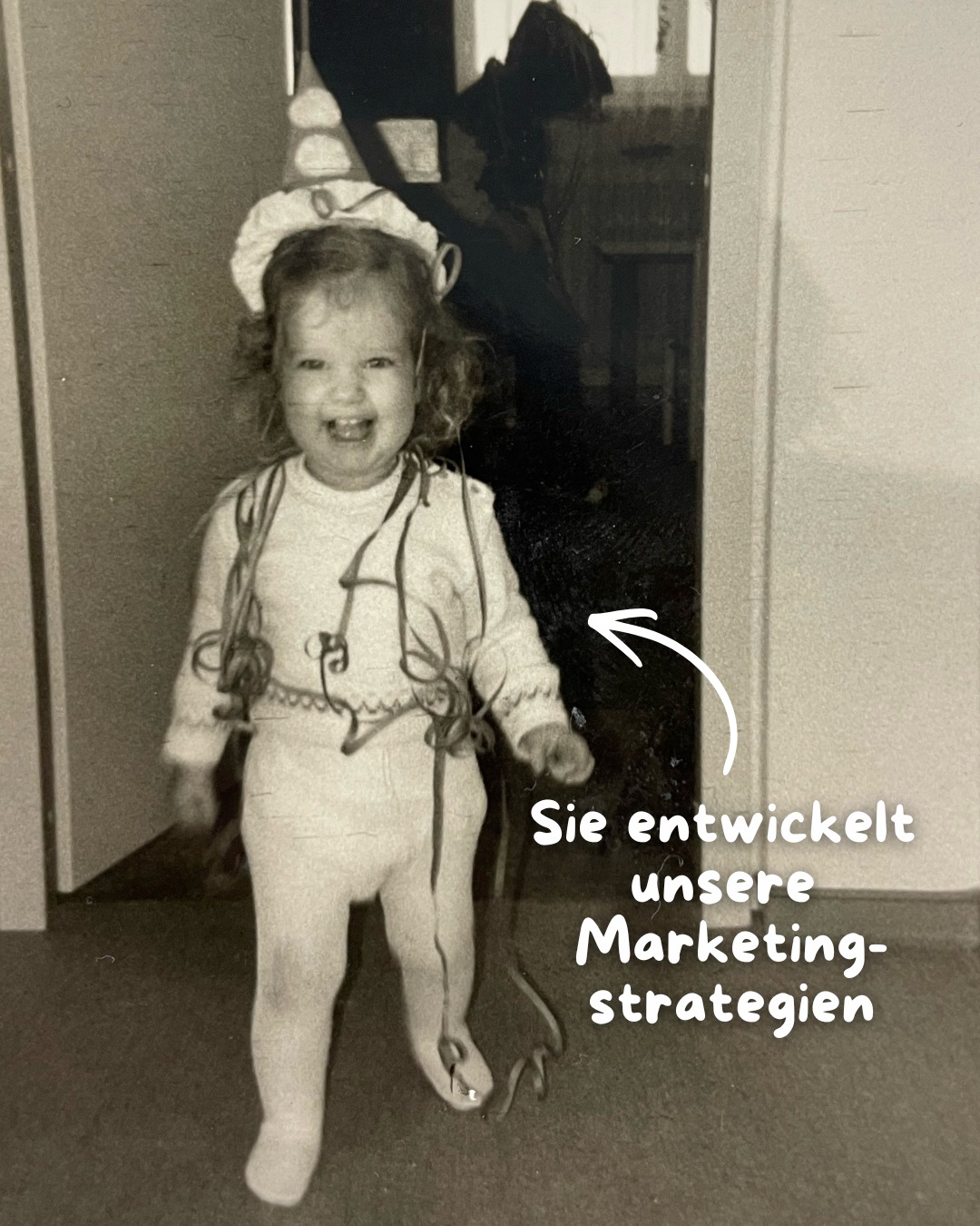 Dieser Alternativtext wurde KI-generiert: Schwarz-weißes Foto eines kleinen Kindes, das lächelnd auf die Kamera schaut. Im Hintergrund ist ein Innenraum zu erkennen.