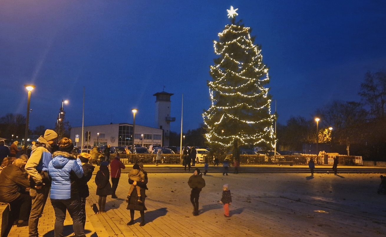 Ein beleuchteter Weihnachtsbaum steht auf einem Platz, umgeben von Menschen. Im Hintergrund sind Gebäude sichtbar.