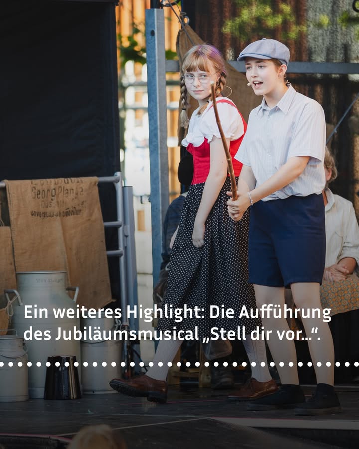 Dieser Alternativtext wurde KI-generiert: Zwei Personen stehen im Freien vor einer verschwommenen Kulisse. Sie scheinen miteinander zu sprechen und tragen Kleidung im Stil vergangener Zeiten.