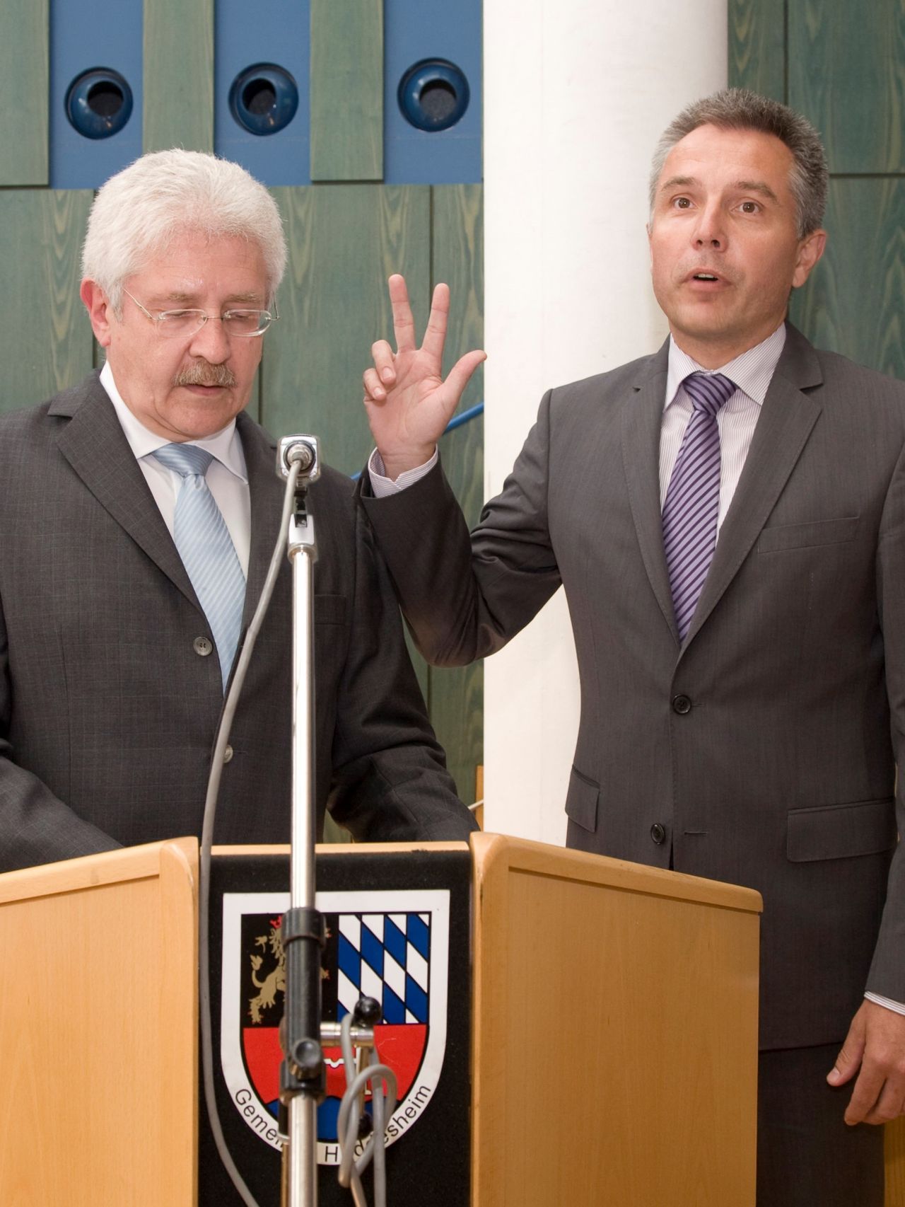 Dieser Alternativtext wurde KI-generiert: Zwei Personen in Anzügen stehen an einem Rednerpult mit Wappen. Eine Person macht eine Handgeste, während die andere zuhört.
