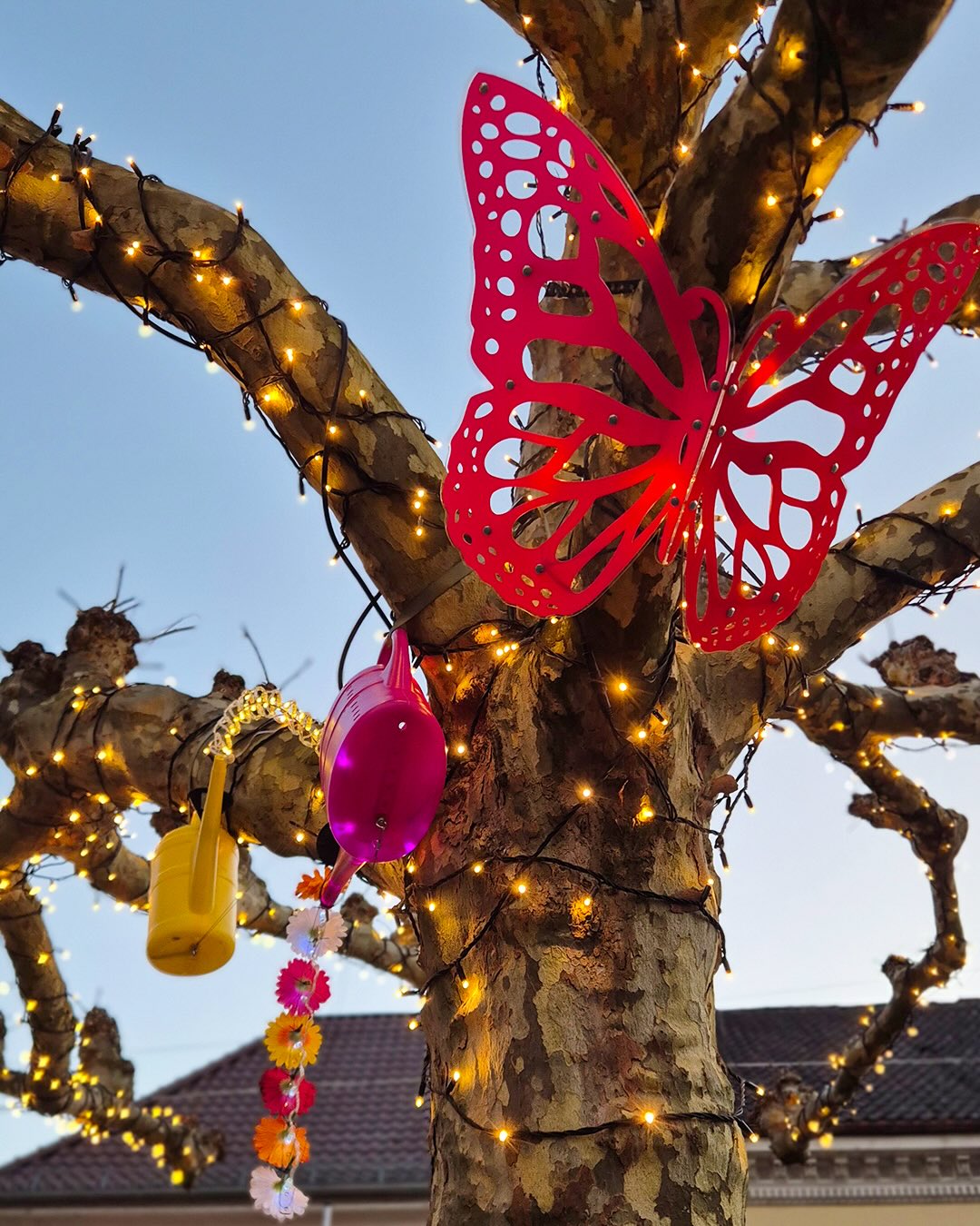 Dieser Alternativtext wurde KI-generiert: Ein Baum ist mit Lichtern und bunten Dekorationsobjekten geschmückt, darunter eine große Schmetterlings- und Blumendekoration. Das Bild wirkt festlich und ist in warmen Farbtönen gehalten.