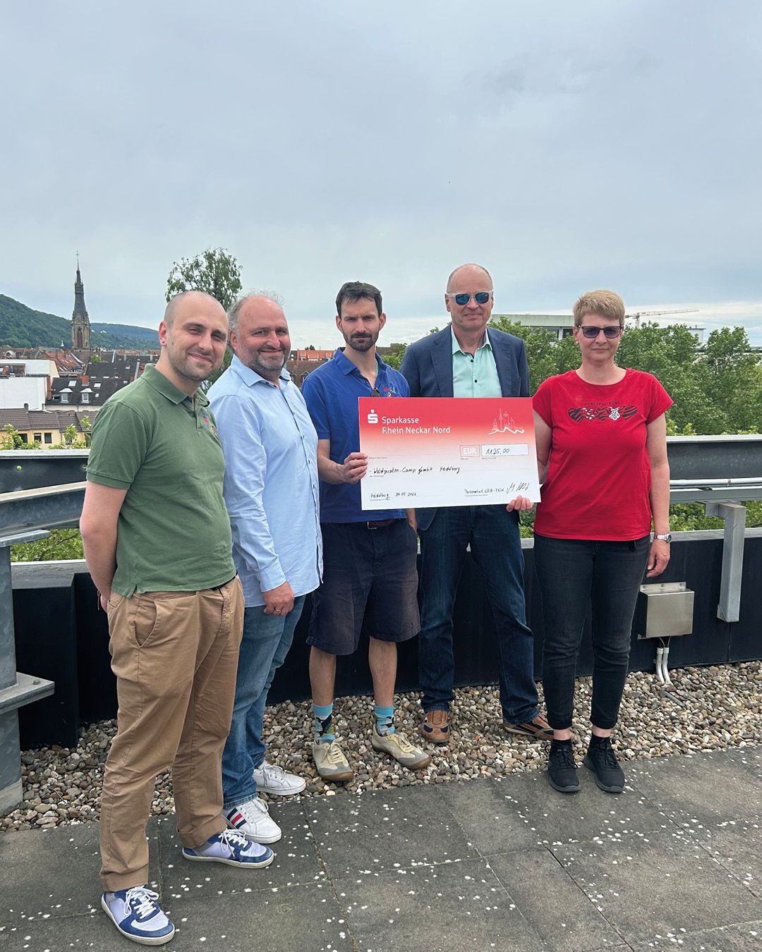 Fünf Personen stehen im Freien auf einer Dachterrasse und halten einen großen Spendenscheck der Sparkasse Rhein Neckar Nord über 1.125,00 EUR für das Waldgarten-Camp Heidelberg. Im Hintergrund sind Gebäude und ein Kirchturm zu sehen. (KI-Alternativtext)