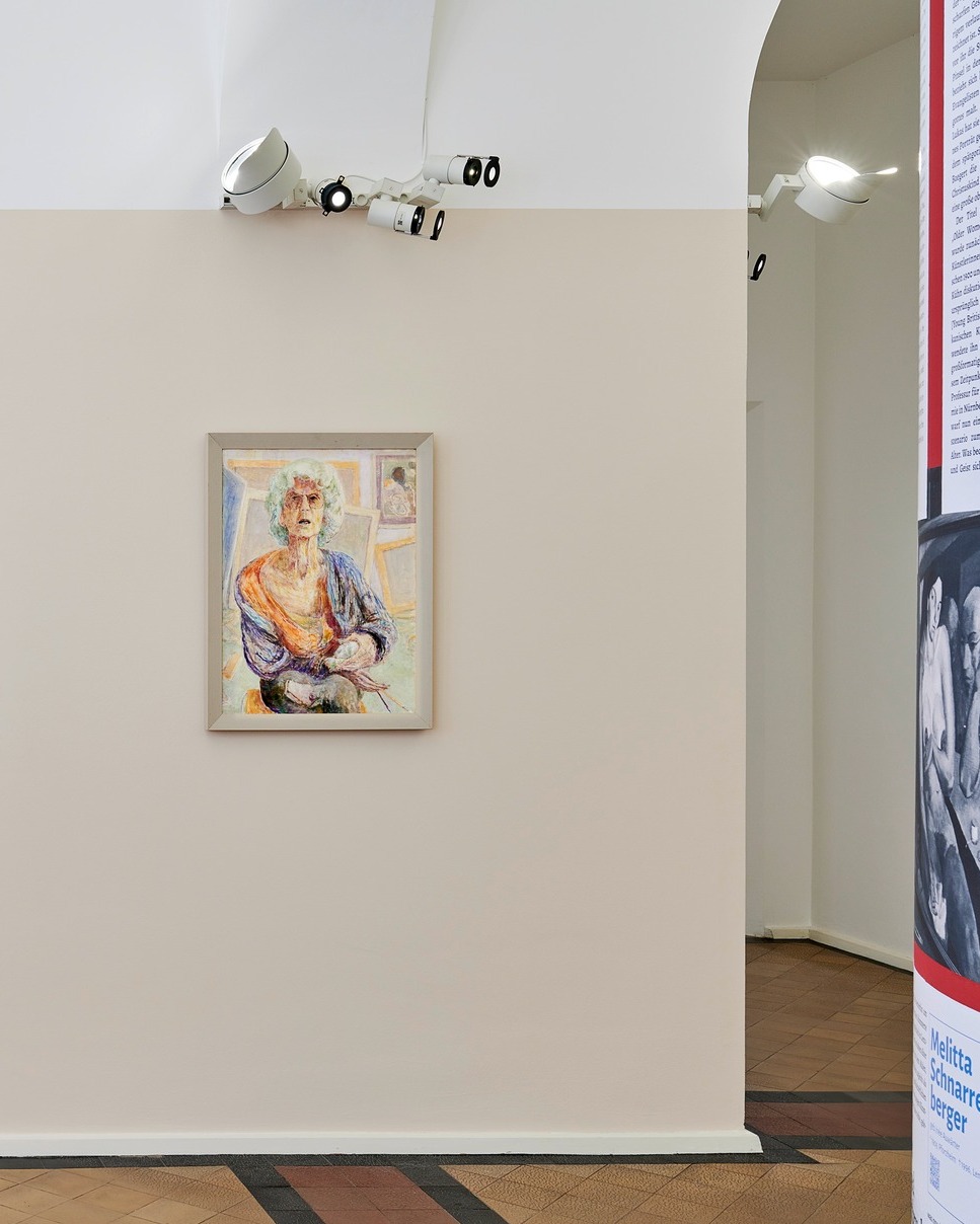 Dieser Alternativtext wurde KI-generiert: Ein Gemälde hängt an einer hellen Wand in einem Raum. Im Hintergrund sind Teile von Texten auf Postern und Deckenleuchten zu sehen.