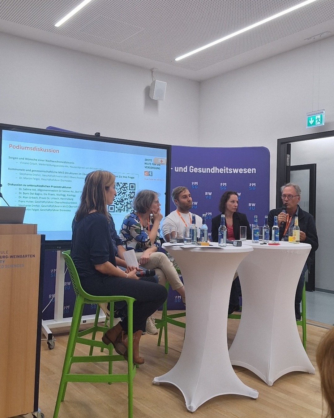 Sechs Menschen sitzen an Tischen nebeneinander und diskutieren bei einer Podiumsdiskussion.