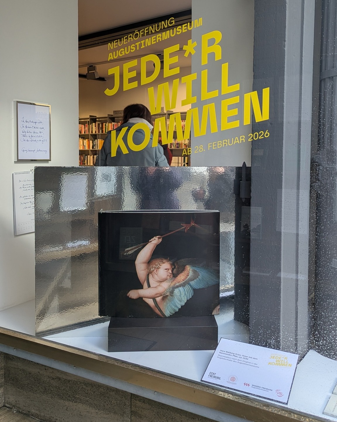 Dieser Alternativtext wurde KI-generiert: Eine Schaufensterdekoration zeigt eine Skulptur und Plakate mit Text. Das Bild ist farbig und durch das Fenster entstanden, was zu Reflexionen führt.