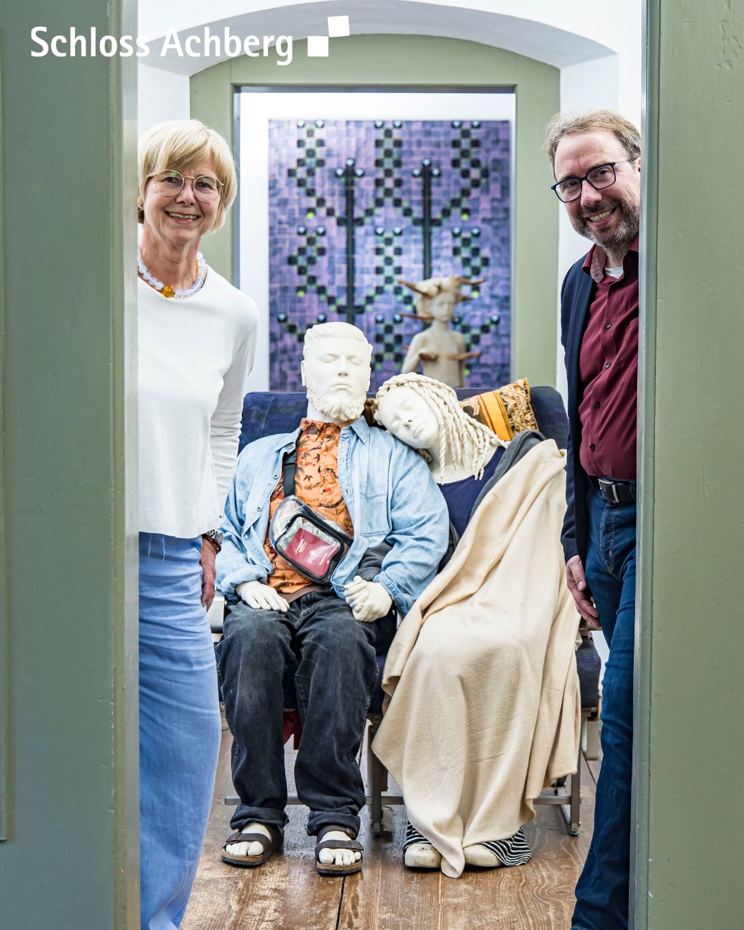 Dieser Alternativtext wurde KI-generiert: Zwei Personen stehen vor einer Kunstinstallation, die eine Sitzgruppe mit einer Büste und weiteren Figuren umfasst. Der Raum wirkt hell und freundlich.