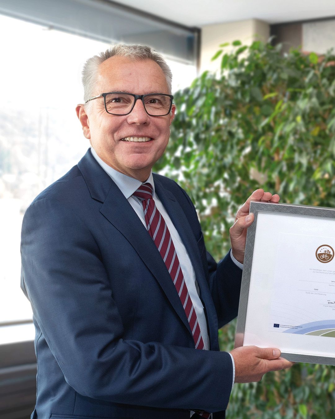 Dieser Alternativtext wurde KI-generiert: Eine Person in einem Anzug hält ein gerahmtes Dokument hoch. Im Hintergrund ist eine Pflanze und ein Blick durch ein Fenster zu sehen.