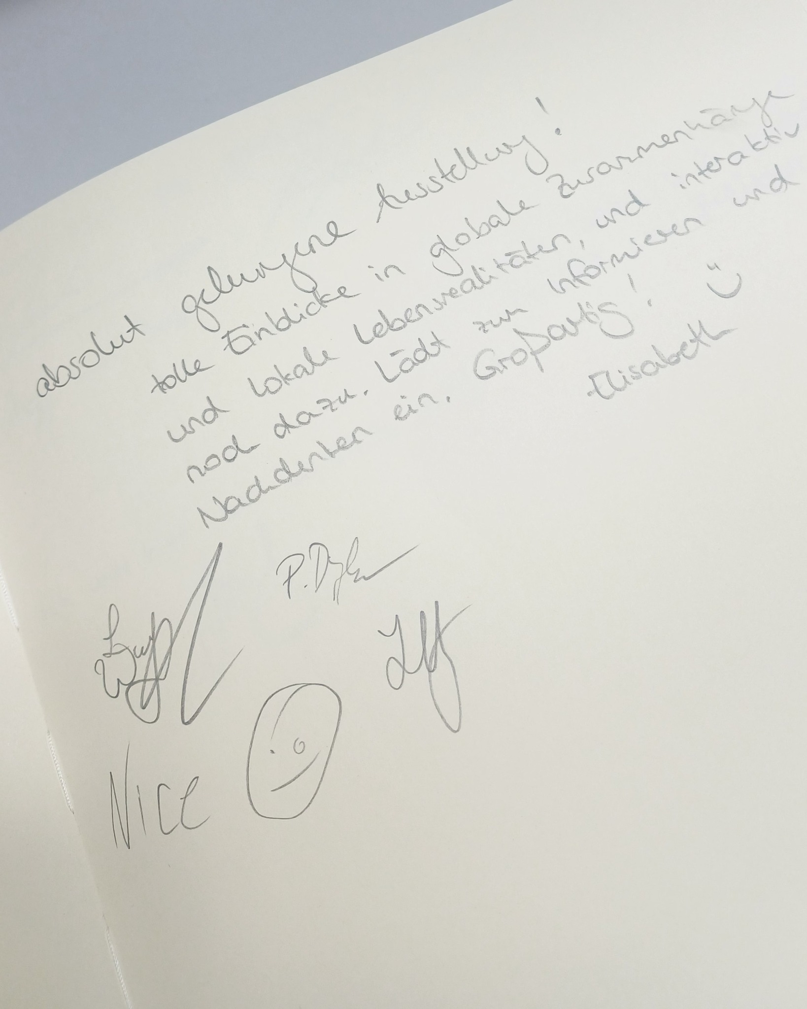 Ein handschriftlicher Text auf weißem Papier lautet: „absolut gelungene Ausstellung! tolle Einblicke in globale Zusammenhänge und lokale Lebensrealitäten, und interaktiv nod dazu. Lädt zum Informieren und Nachdenken ein. Großartig! :) -Elisabeth“. Darunter sind mehrere Unterschriften, das Wort „Nice“ und ein gezeichnetes Smiley zu sehen. (KI-Alternativtext)