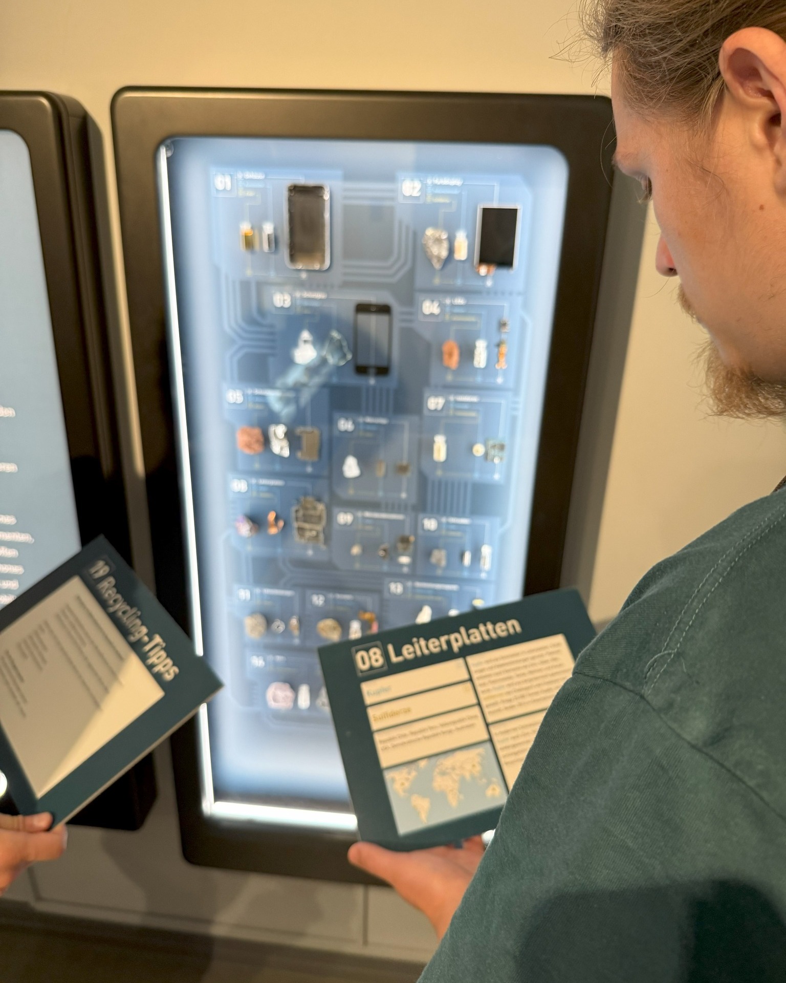 Dieser Alternativtext wurde KI-generiert: Eine Person betrachtet eine beleuchtete Vitrine mit verschiedenen elektronischen Komponenten und Platinen. Daneben liegen Informationsblätter mit Beschriftungen.