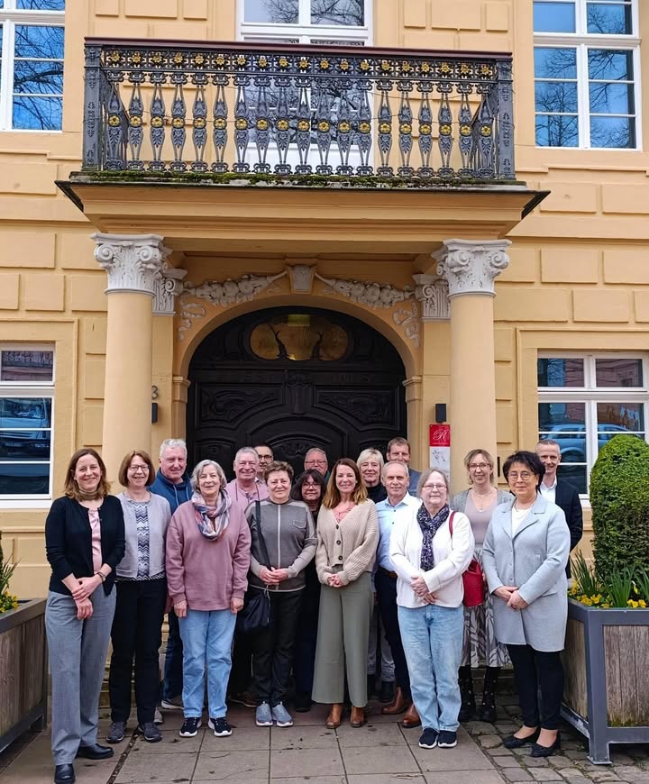 Dieser Alternativtext wurde KI-generiert: Ein gelbes Gebäude mit einem verzierten Balkon und einem dunklen Eingangsbereich. Eine Gruppe von Menschen steht vor dem Gebäude und posiert für ein Foto.