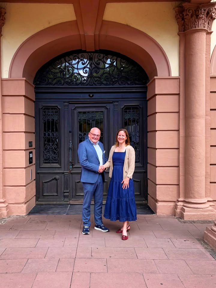 Zwei Personen stehen Hand in Hand vor einem großen, dunklen Eingang unter einem Bogen. Die Person links trägt einen blauen Anzug, die Person rechts ein langes blaues Kleid mit einer hellen Jacke. Beide blicken lächelnd in die Kamera. (KI-Alternativtext)