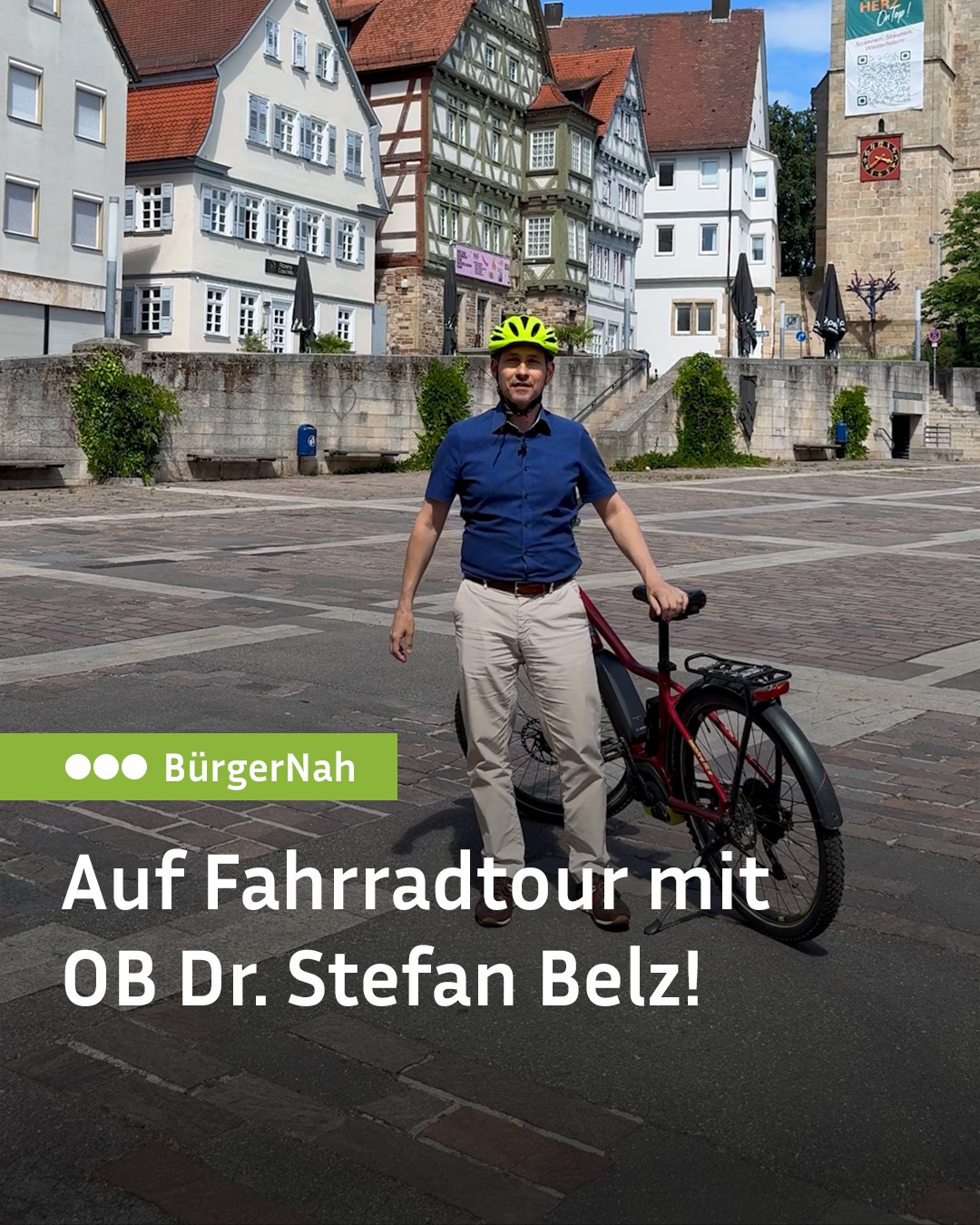 Eine Person mit gelbem Helm, blauem Hemd und beigen Hosen steht mit einem roten E-Bike auf einem Platz vor historischen Fachwerkhäusern. Ein grüner Balken zeigt das Logo „BürgerNah“ und darunter steht der Text „Auf Fahrradtour mit OB Dr. Stefan Belz!“. (KI-Alternativtext)